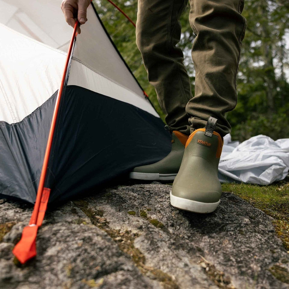 Olive green XTRATUF Ankle Deck Camp Boots with orange trim, worn by a person setting up a tent.