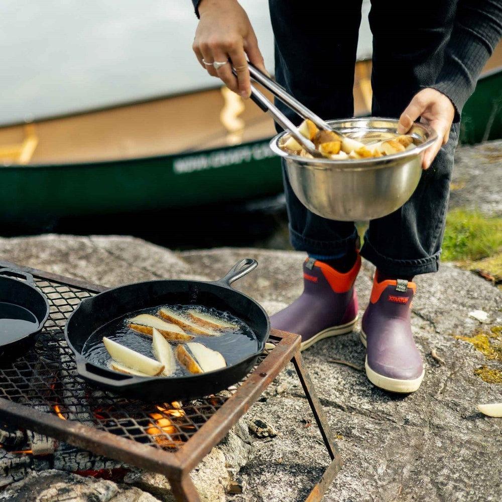 Person in black pants and purple XTRATUF Ankle Deck Boots frying potatoes outdoors.