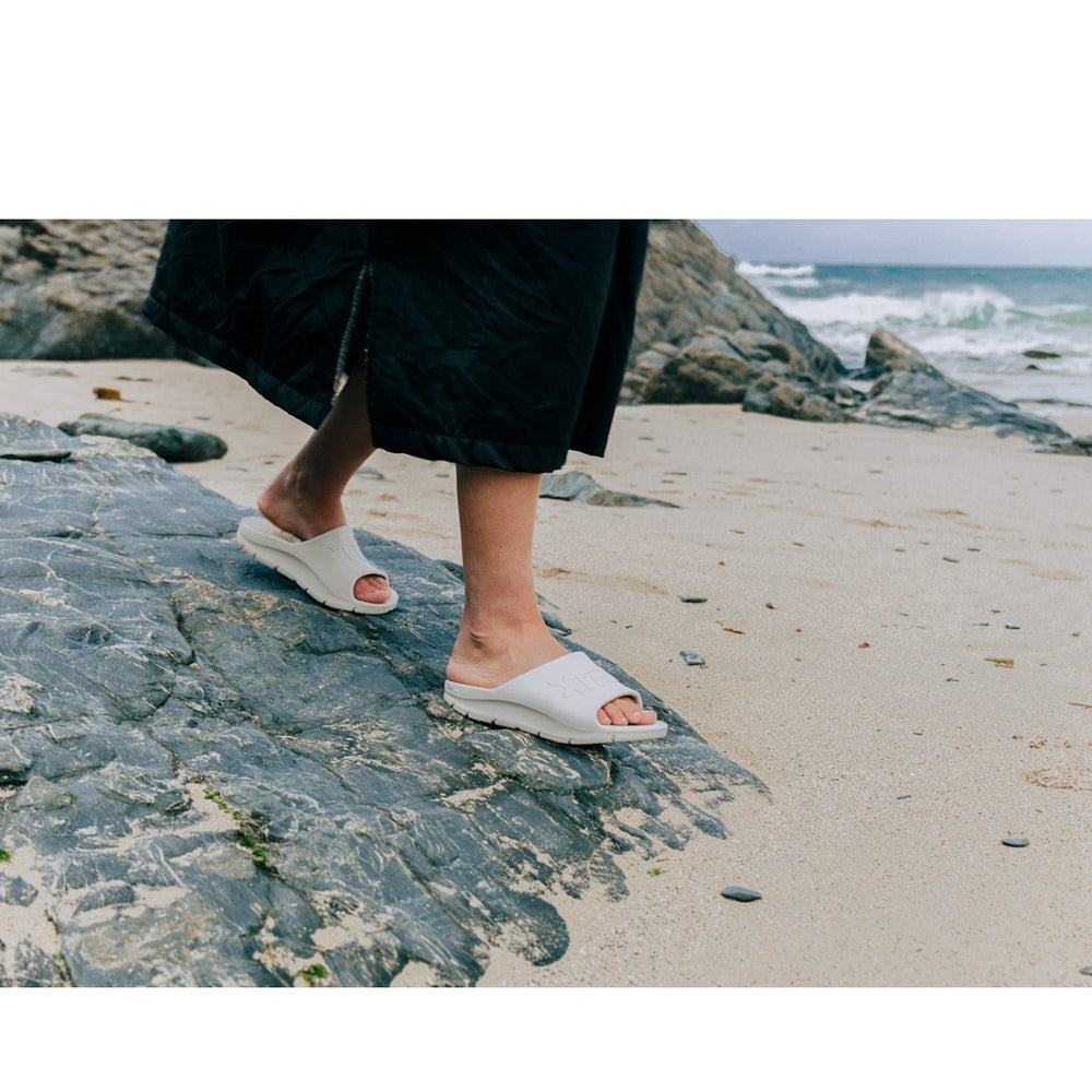 White Apres Fish slide sandals on feet walking on a rocky beach.