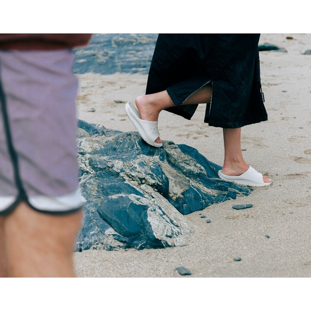 White Apres Fish slide sandals on a person's feet, walking on a sandy beach with dark rocks.
