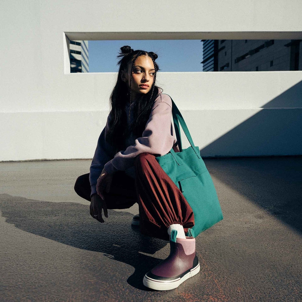 Woman in a lavender sweatshirt and maroon pants, crouching with a teal tote bag and wearing purple Ankle Deck Boots.