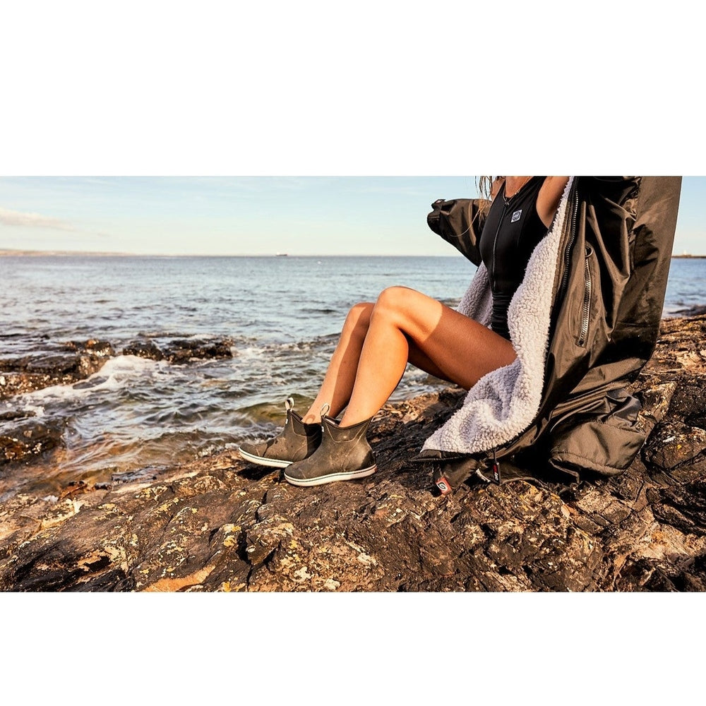 Woman wearing black Xtratuf Ankle Deck Boots and a black robe, sitting on rocks by the ocean.