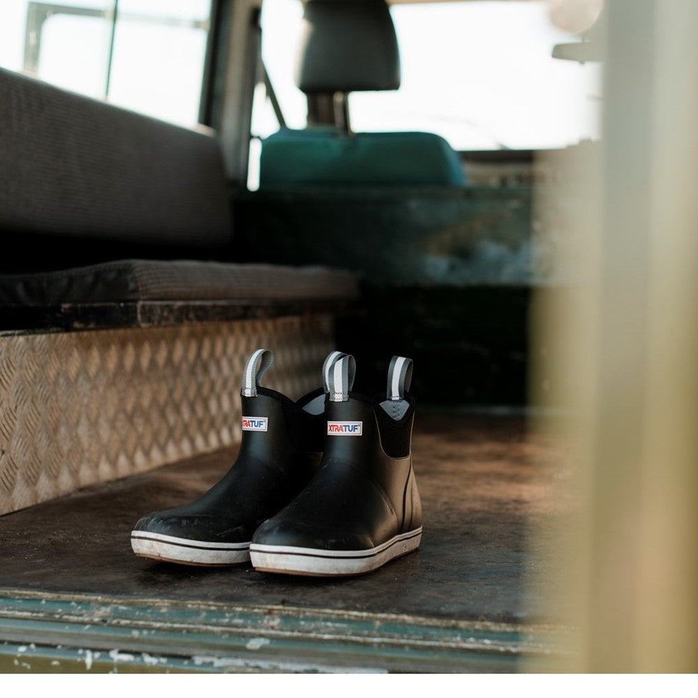 Pair of black Xtratuf women's ankle deck boots with white soles and pull tabs, on a textured metal floor.