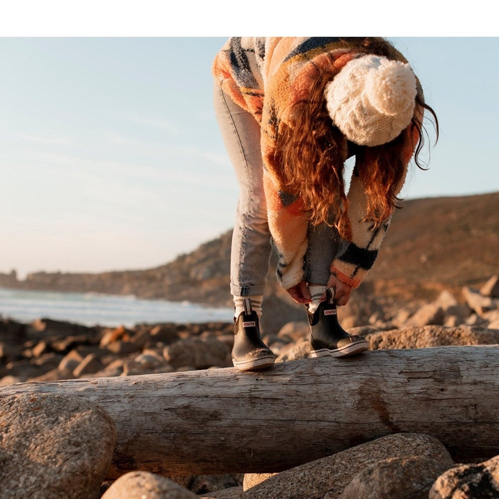 Woman in a colorful sweater and jeans adjusting black Xtratuf Ankle Deck Boots on a log by the ocean.