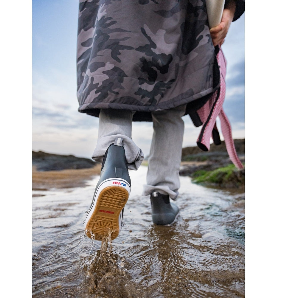 Black Xtratuf Ankle Deck Boots with slip-resistant Chevron outsole, worn by a person walking through shallow water.