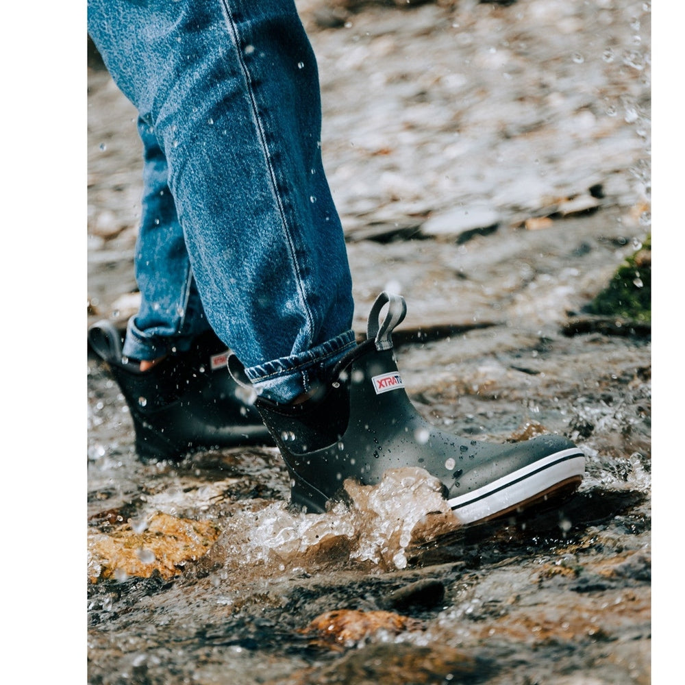 Black Xtratuf Ankle Deck Boots with white trim, worn by a person in jeans, splashing through water on rocks.